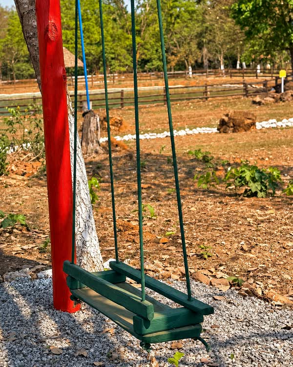 Balanço de assento verde preso a um poste vermelho, em terreno de brita